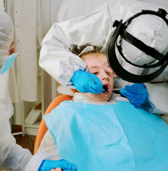Dental professional examining a young patient’s mouth while wearing protective equipment